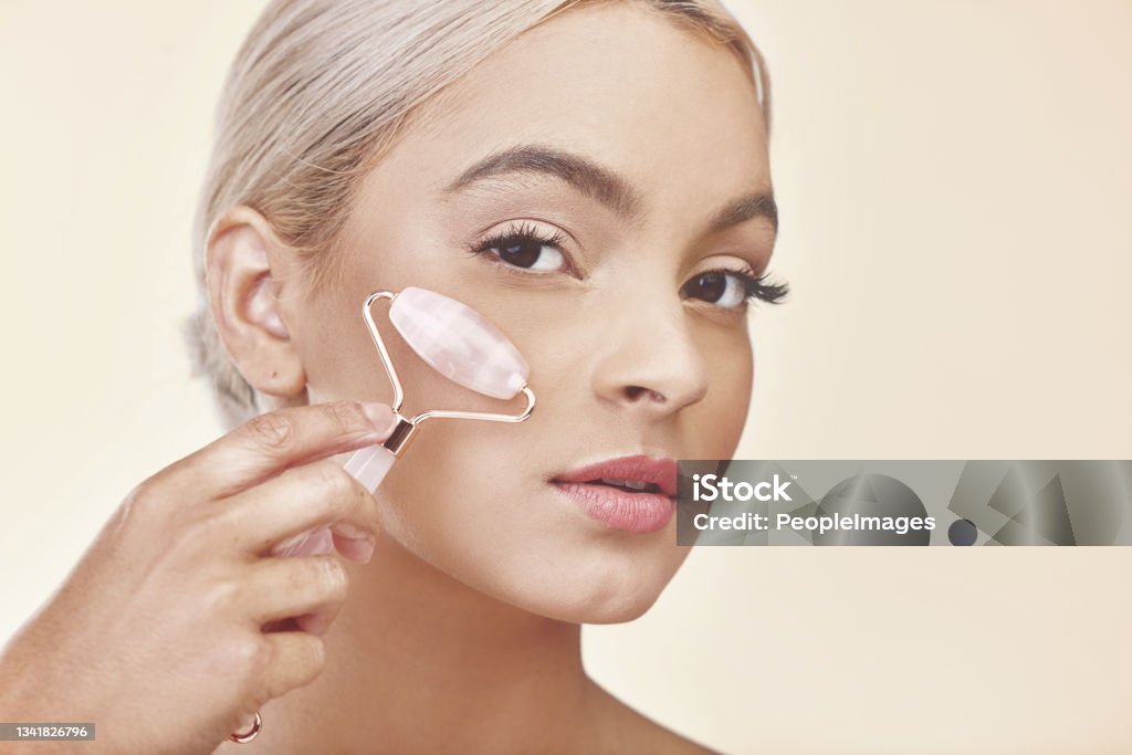 Studio shot of a young woman using a jade roller on her face A good skincare regime is easily noticeable Jade Roller Stock Photo