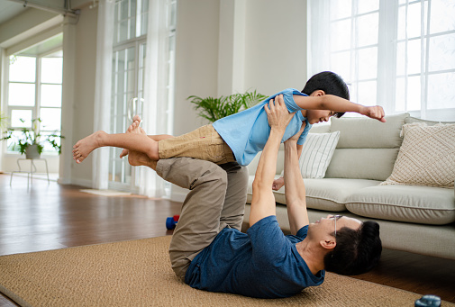 Asian father lying on carpet floor, lifting excited happy son at home.
