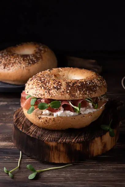 Ham bagel for breakfast on a dark wooden background, sandwich with ricotta, prosciutto and microgreens, rustic style.