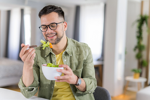 Happy young man eating a healthy salad for breakfast in the living room.