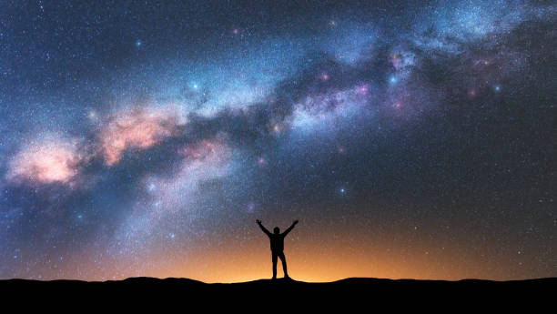 milky way and happy man at night. silhouette of alone guy with raised up arms, sky with bright stars, orange light in summer. galaxy. space background. landscape with milky way. travel and nature - milky way galaxy space star imagens e fotografias de stock