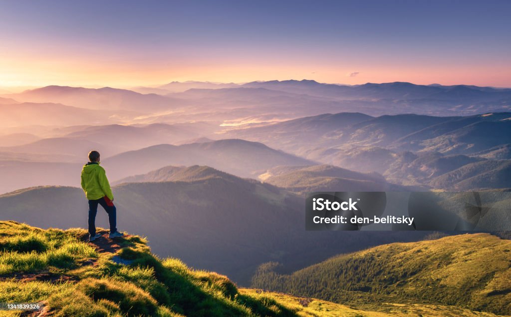 Sporty man on the mountain peak looking on mountain valley with sunbeams at colorful sunset in autumn in Europe. Landscape with traveler, foggy hills, forest in fall, amazing sky and sunlight in fall Men Stock Photo