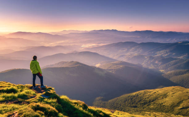 sporty man on the mountain peak looking on mountain valley with sunbeams at colorful sunset in autumn in europe. landscape with traveler, foggy hills, forest in fall, amazing sky and sunlight in fall - mountain peak imagens e fotografias de stock