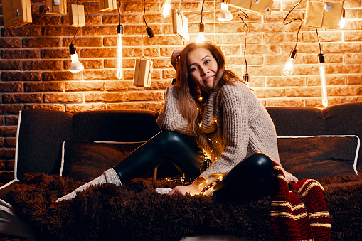 Pretty girl in sweater sitting on couch. Christmas and New Year atmosphere. Different shapes light bulbs on background of brick wall. Striped red scarf on sofa. Nice woman on fluffy blanket.