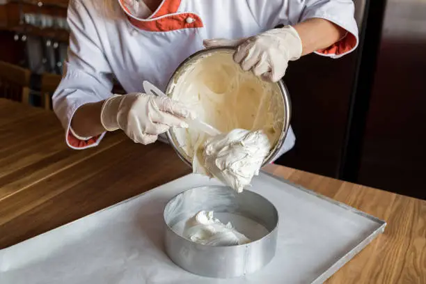 Female chef cook in rubber gloves pours the vanilla cake-mix dough into form for making cake. Young woman baker pouring the dough into baking dish