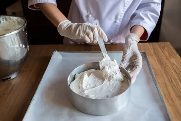 Female chef cook in rubber gloves preparing vanilla cake-mix dough in form for baking cake. Young woman baker pouring the dough in baking dish