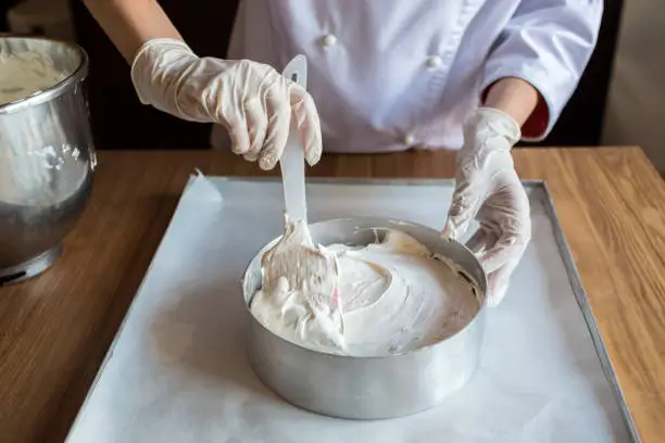 Female chef cook in rubber gloves preparing vanilla cake-mix dough in form for baking cake. Young woman baker pouring the dough in baking dish