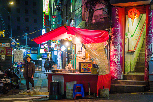 Pedestrians passing food stall illuminated at night on the neon lit streets of central Seoul, South Korea.