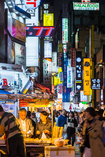 People buying food from a stall at night in the neon lit shopping streets of Myeong-dong in central Seoul, South Korea.