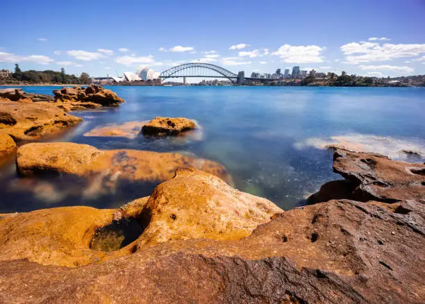 Photo of Sydney Harbour with nice rocks in the foreground the soft waves crashing on the shore and the beautiful harbour foreshore as a backdrop NSW Australia
