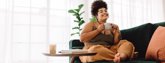 Happy female holding coffee mug while sitting on couch at home. Smiling woman sitting on sofa having coffee.
