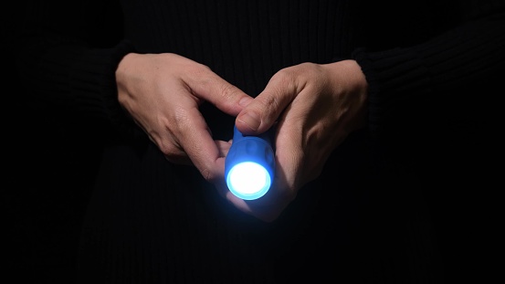 Hand with a led flashlight shining in the darkness with blue beam into the camera, spy and investigation concept.