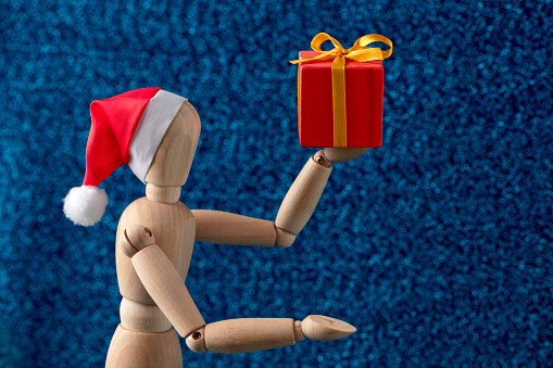 Close-up of a wooden mannequin in Santa hat holding a gift box and showing something by the hand. Blue defocused background, space for your Christmas message.