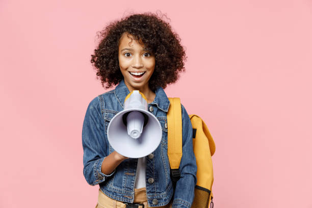 leader sorridente piccola ragazza afroamericana bambina scuola 12-13 anni in abiti casual denim con zaino urlo in megafono isolato su sfondo rosa pastello studio concetto di educazione infantile. - 12 13 years immagine foto e immagini stock