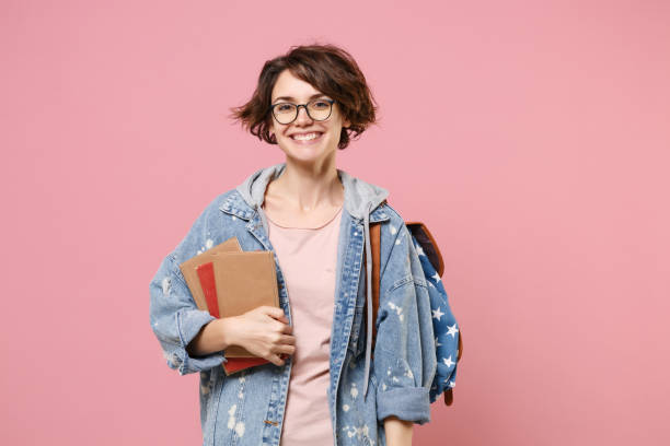 istock Smiling young woman student in denim clothes, eyeglasses, backpack posing isolated on pastel pink background. Education in high school university college concept. Mock up copy space. Holding books. 1341906169