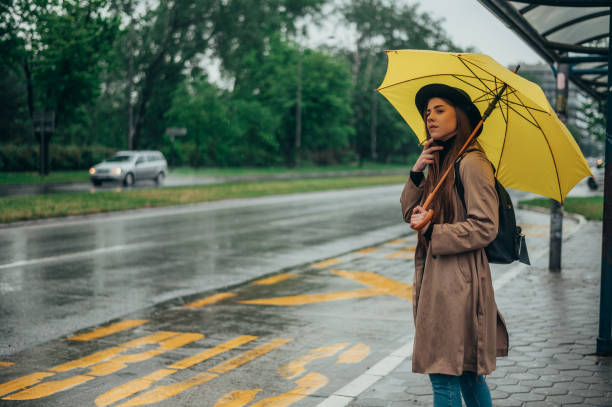 istock Woman holding a yellow umbrella and standing on a bus stop 1341908146