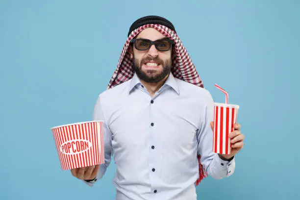 Confused arabian muslim man in keffiyeh kafiya ring igal agal 3d imax glasses isolated on pastel blue background. People religious concept. Watching movie film, holding bucket of popcorn, cup of soda