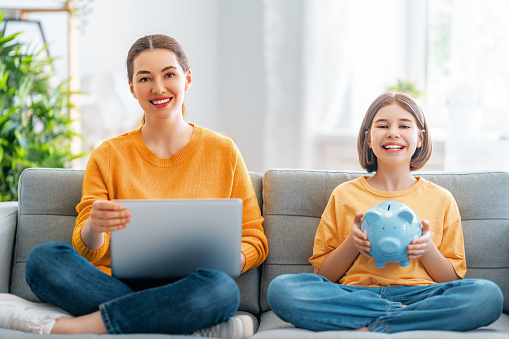 Mother and daughter are holding piggy bank and laptop sitting on the sofa at home.
