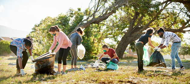 Diverse group of young volunteers wearing face masks collecting litter in bags during a community cleanup in a nature reserve