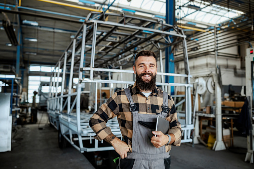 A happy auto-industry worker standing with a tablet under the armpit and looking at the camera. Auto-industry worker at the factory