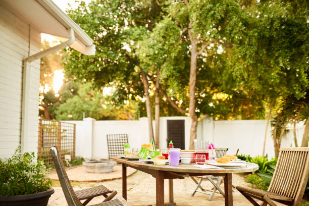 Large wooden table with rustic chairs set up for an outdoor meal in a lush green Swedish garden. Decorated with fresh flowers and clear glassware. A perfect setting for summer feasts, countryside living, or farm-to-table themes.