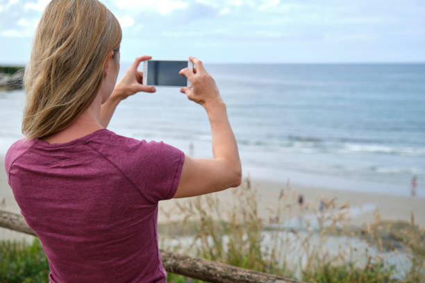 istock Woman taking photograph with smart phone camera on the beach, Back view. 1341913432