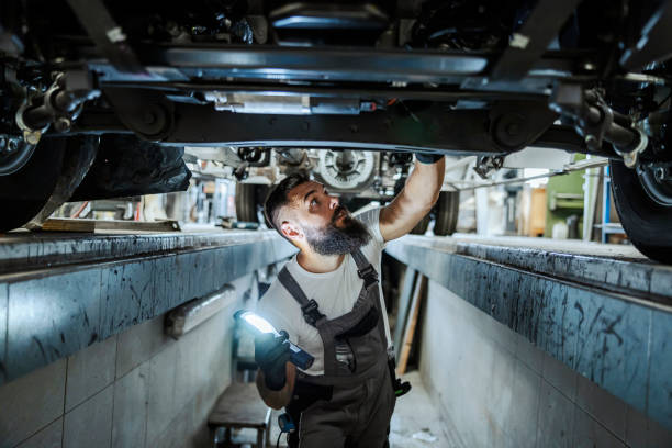 An auto-mechanic is standing in a car mechanic's pit and looking car bottom. He is lightning and looking for malfunction. A worker in the car repair workshop. stock photo