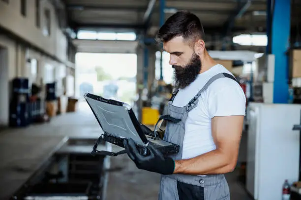 An auto-mechanic is standing in his workshop and using the special laptop for car maintenance. Worker analyzing malfunction on vehicles. Worker in workshop