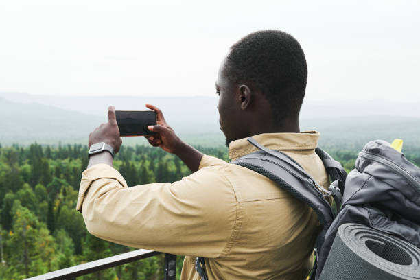 istock Young African American man with backpack standing on bridge and photographing forest landscape on phone 1341928439