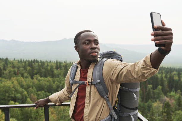 istock Handsome young Black man with backpack standing on bridge and taking selfie against forest landscape 1341928557