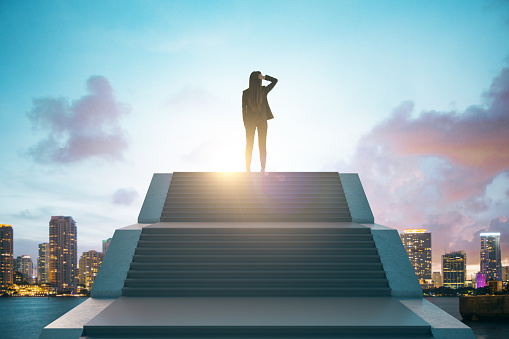 Back view of young european businesswoman standing on top of stairs on bright city background with sky view. Leadership, growth and success concept