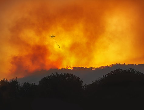 Helicopter with water bucket fighting forest wildfire at night, dramatic landscape with red sky and heavy smoke