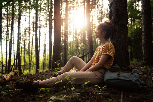 Cheerful young Caucasian woman sitting on the ground in the shade of a pine tree and resting after a long hike.