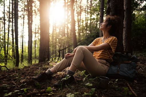 Cheerful young Caucasian woman sitting on the ground in the shade of a pine tree and resting after a long hike.