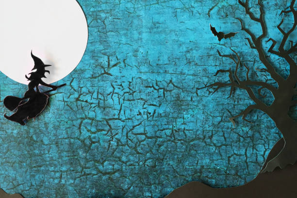 Stock photo showing a Halloween night scene poster / background design with homemade cut out full moon, bat, witch flying on broomstick and bare tree shapes on cracked blue background with copy space.