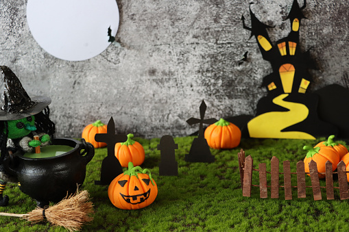 Stock photo showing a Halloween night scene of cemetery with full moon, homemade cut out haunted house, bats, deciduous tree, pumpkin Jack O'lanterns, string witch stirring cauldron, broom and tombstones on artificial grass surface against a black background.