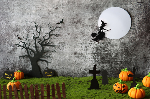 Stock photo showing a Halloween night scene of cemetery with homemade cut out full moon, bat, deciduous tree, pumpkin Jack O'lanterns, witch flying on broomstick and tombstones on artificial grass surface against a black background.
