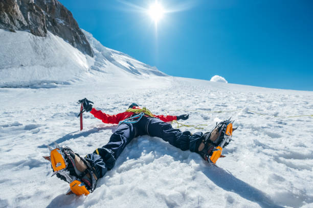 Tired exhausted climber lying under Mont Blanc du Tacul mountain. Wide opened legs in boots with crampons and mountaineering clothes with blue sky with bright sun background. Active people concept. stock photo