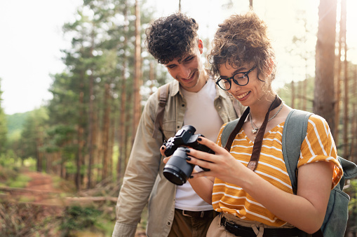 Cheerful young Caucasian female photographer and her boyfriend looking at the photos on the camera while hiking in the woods together. Traveling concept.