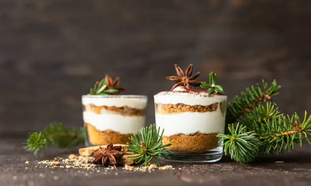 Layered dessert in glass jar with cookie crumble and whipped cream decorated with rosemary and anise, dark brown background. No bake cheesecake, trifle or pudding.