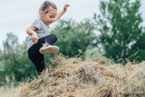 petite fille rieuse de 3-4 ans saute et joue dans une botte de foin, sur fond d’arbres - child caucasian little girls 3 4 years photos et images de collection