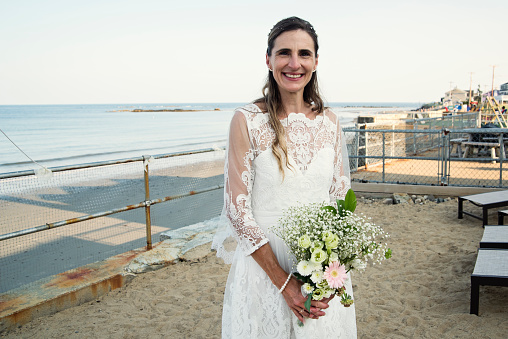 Mature bride portrait at beach house. She is in her forties, and is looking at the camera with a smile. Ocean in the background. Horizontal waist up outdoors shot with copy space.
