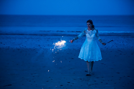 Mature bride lighting bengal fire on the beach at dusk. Bride is in her forties, and wearing her wedding dress. Horizontal full length outdoors shot with copy space.
