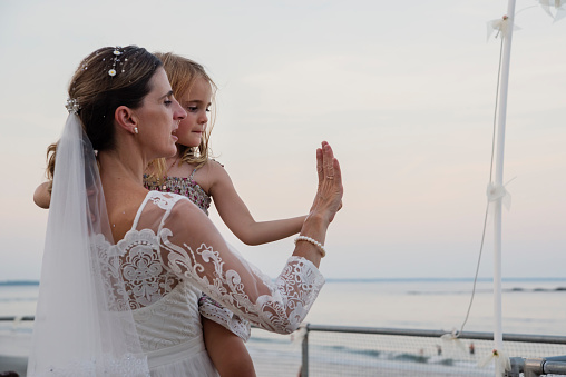 Bride dancing with her little daughter in floral dresses near the ocean. She is in her forties and daughter is 4 year’s old. Horizontal waist up outdoors shot with copy space.