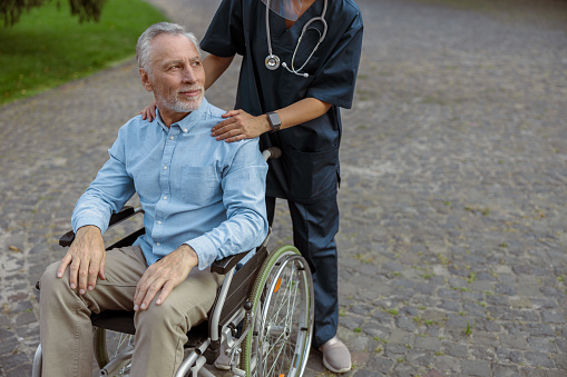 Recovering male patient in a wheelchair looking away on a walk with a nurse outdoors. Medical insurance, rehabilitation concept