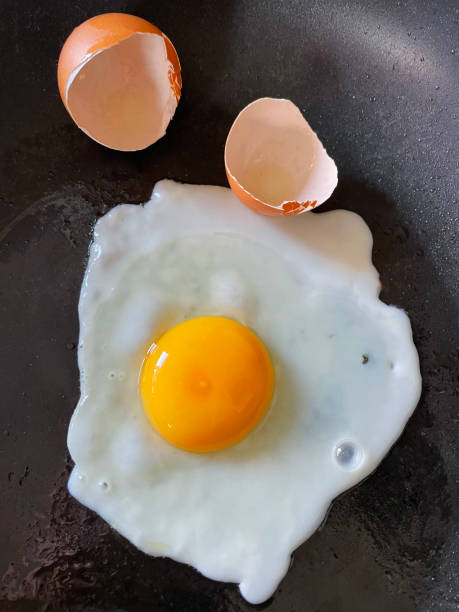 istock Close-up image of sunny side up fried egg sat seasoned in non-stick frying pan, egg shell halves, elevated view 1341951450