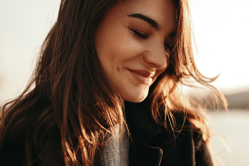 Attractive young long haired brunette with eyes closed smiling happily while enjoying fresh wind and sunlight in autumn nature