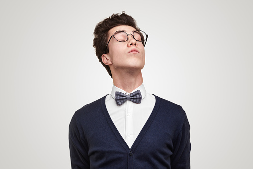Snobbish young man in glasses closing eyes and raising head against gray background