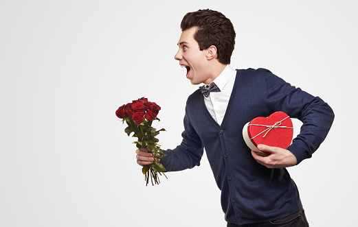 Side view of cheerful young romantic guy in elegant outfit carrying bouquet of red roses and heart shaped gift box, while rushing to date on white background
