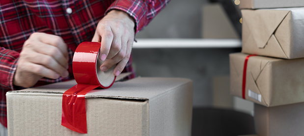 Close-up of man's hands in a red plaid shirt, sealing the box with red tape. Christmas sale concept.